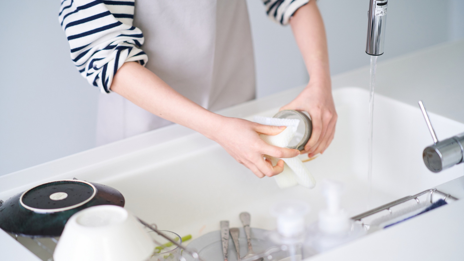 A person in a striped shirt and apron hand-washes a glass under running water in a white sink.