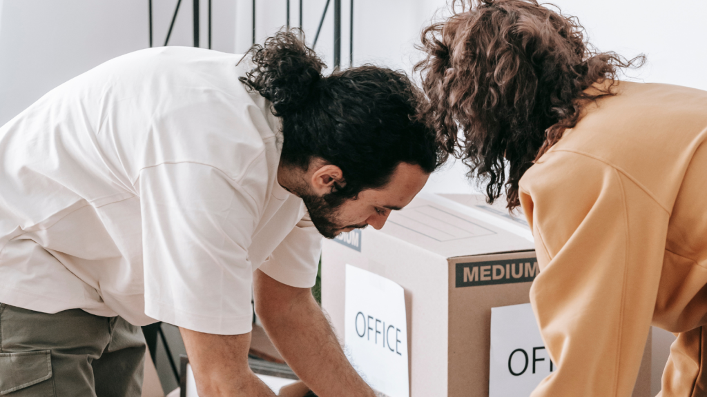 Two people lean over large cardboard boxes labeled “office,” organizing or packing items in a bright room.