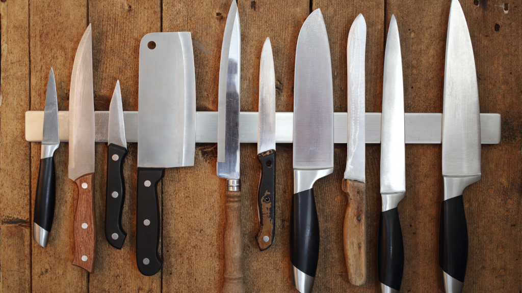 A row of kitchen knives with mixed handles neatly displayed on a magnetic strip against a wooden wall.