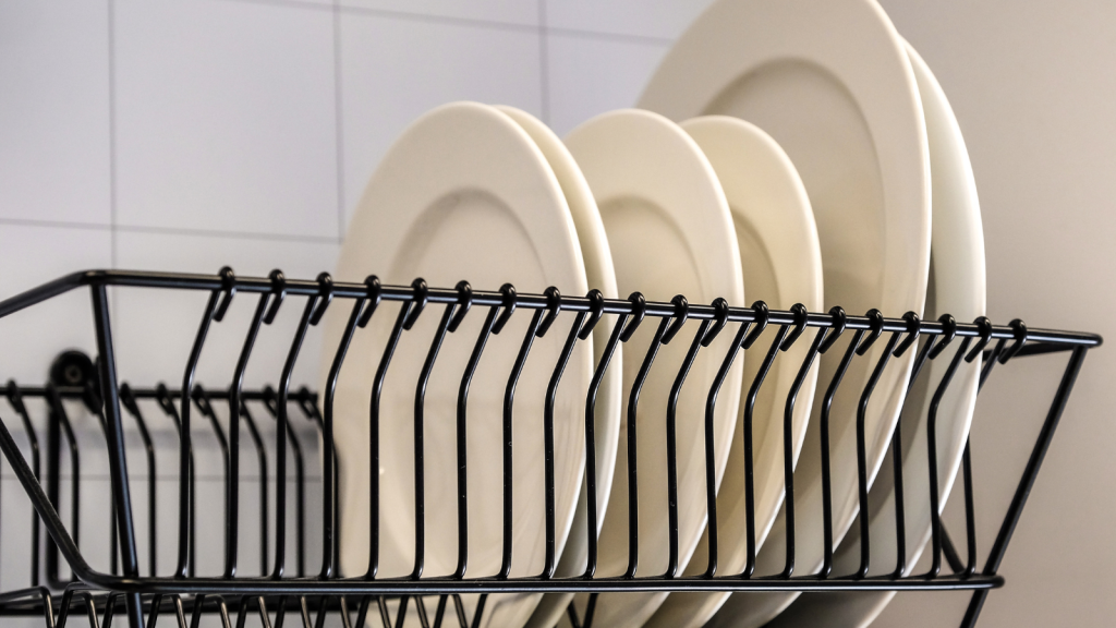 A close-up of white plates drying in a black wire dish rack against a tiled kitchen wall.