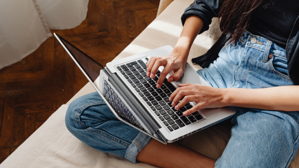 A person in jeans sits cross-legged on a couch, typing on a laptop with natural light coming through a nearby window.