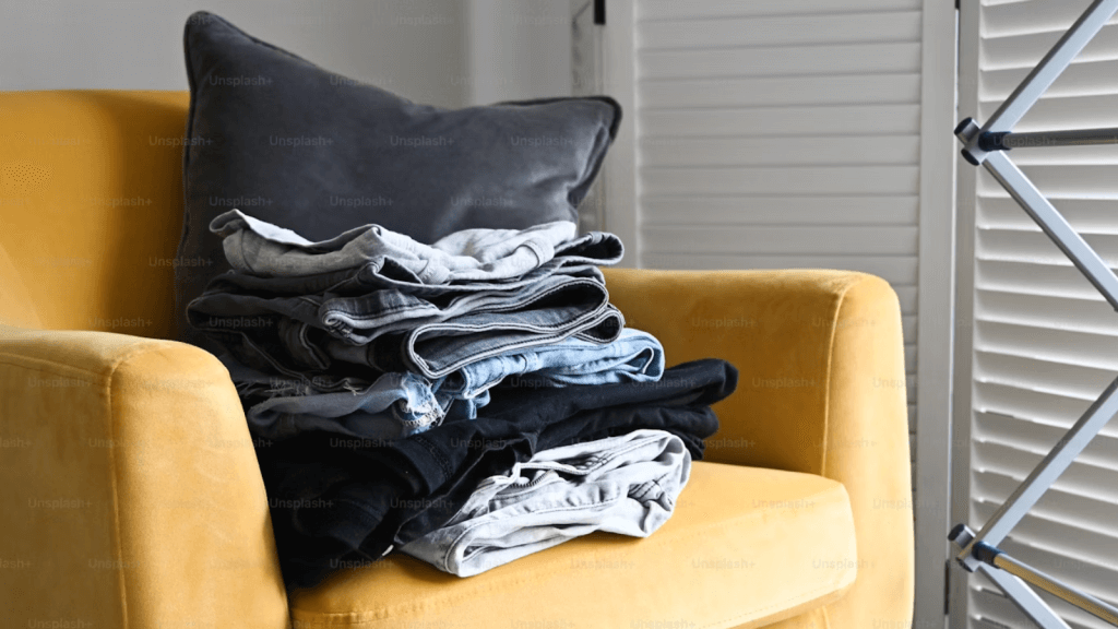 A neat stack of folded jeans and pants on a mustard-yellow chair with a dark gray pillow beside an indoor drying rack.