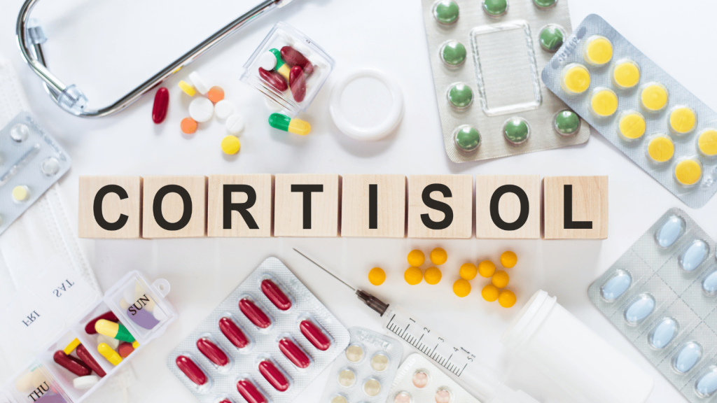 Wooden letter blocks spelling “CORTISOL” sit centered on a table surrounded by various medications, including pills, capsules, blister packs, a syringe, and a stethoscope.
