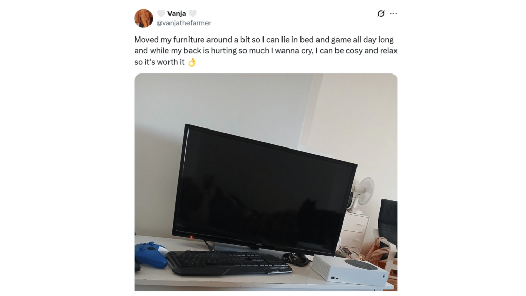 Screenshot of an X post from “Vanja” (@vanjathefarmer) saying she moved furniture so she can lie in bed and game all day; her back hurts but it’s worth it to be cozy and relaxed. The photo shows a tilted TV or monitor on a white desk with a keyboard, mouse, game controllers, and a small console, against a plain wall.