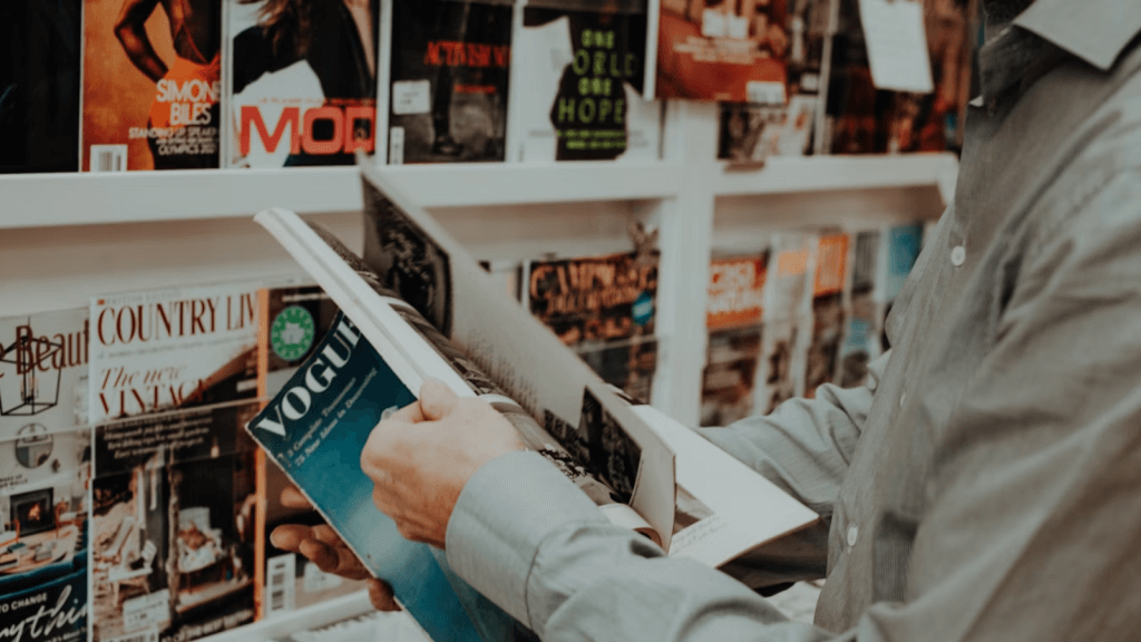 A person in a gray shirt flips through a Vogue magazine in front of a display of various lifestyle and fashion publications.