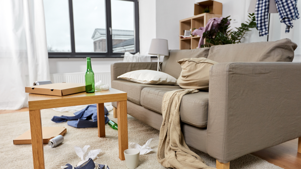 A living room with a beige sofa, coffee table, pizza boxes, and scattered clothes and bottles on the floor.