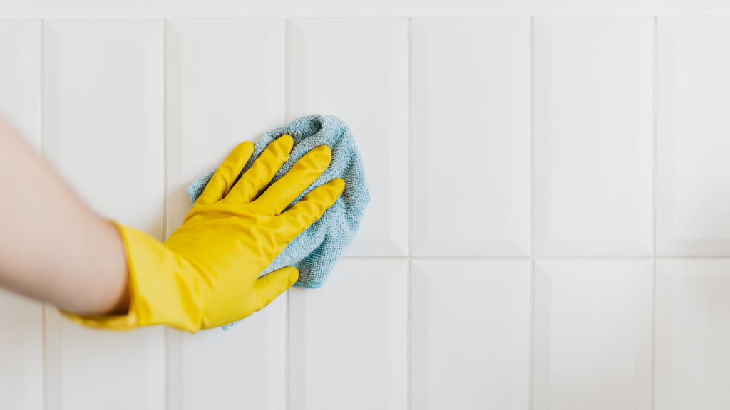 A person wearing yellow gloves wipes white tiled walls with a blue microfiber cloth.