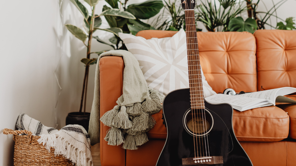A black acoustic guitar leans against a tan leather couch with a blanket and pillow, surrounded by green plants.