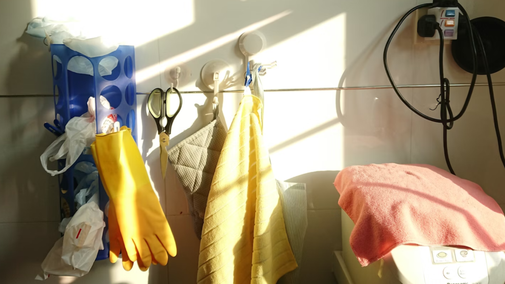 A bright laundry area with yellow gloves, hanging towels, and cleaning tools illuminated by warm sunlight.