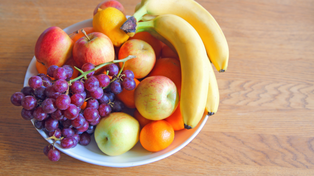 A bowl of apples, bananas, grapes, and citrus fruit sitting on a wooden tabletop.