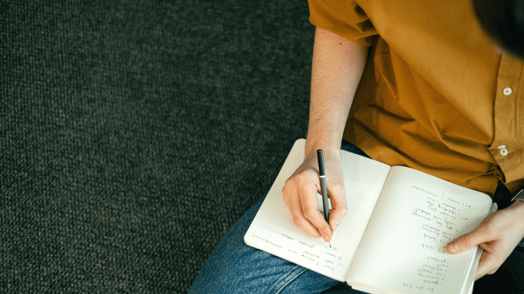 A person sits on the floor writing in an open notebook filled with handwritten notes. Only their torso, arm, and hand are visible, and the background is a textured dark carpet.
