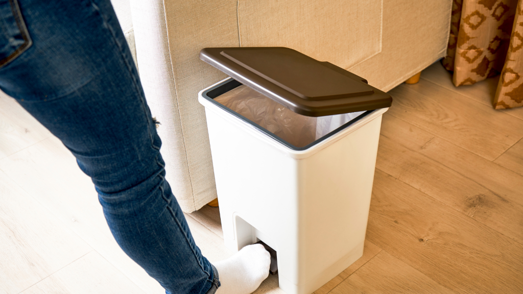 A person’s foot presses the pedal of a white trash bin beside a beige couch.