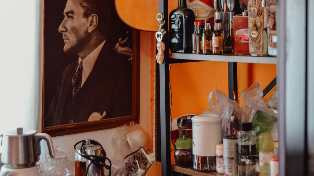 A metal shelf filled with jars, bottles, and knickknacks beside an orange wall and a framed portrait.