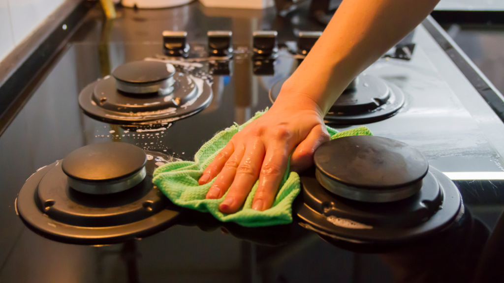 A hand wipes a black gas stovetop with a green cloth, removing soap and water.
