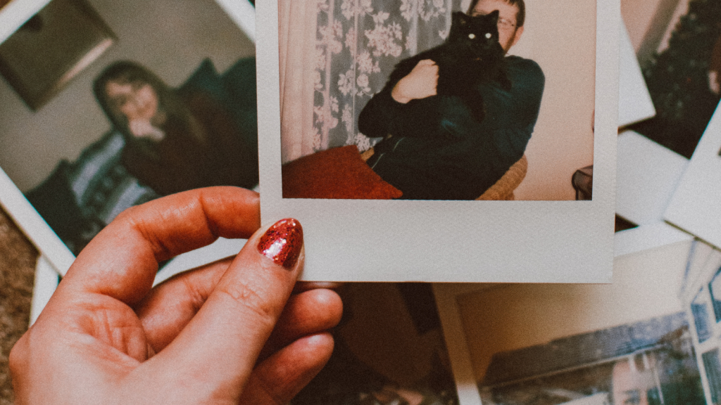A hand with red glitter nail polish holds a Polaroid photo over a table scattered with other old photos.