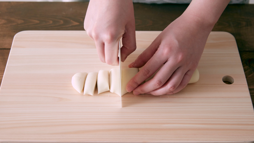 Hands use a knife to cut sections of soft dough on a pale wooden cutting board.