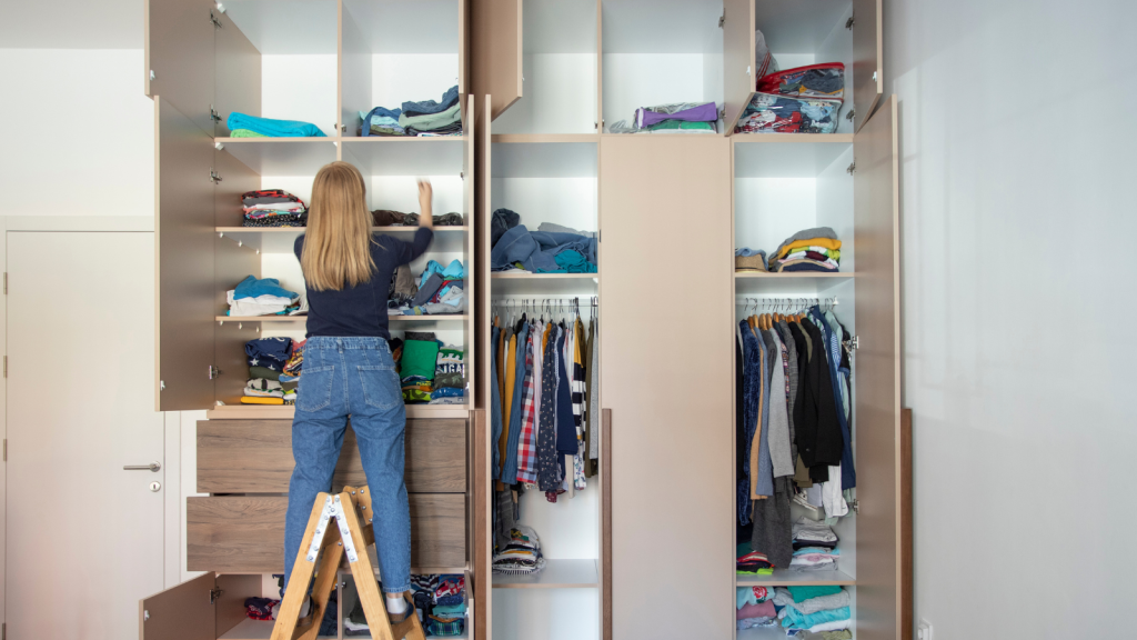 A woman on a small step ladder arranges folded clothes in a tall open wardrobe filled with shirts, sweaters, and hanging items.