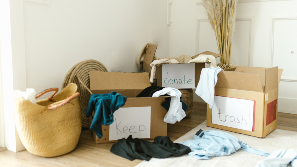 Three cardboard boxes labeled “keep,” “donate,” and “trash” sit on the floor surrounded by piles of clothes and a woven basket.