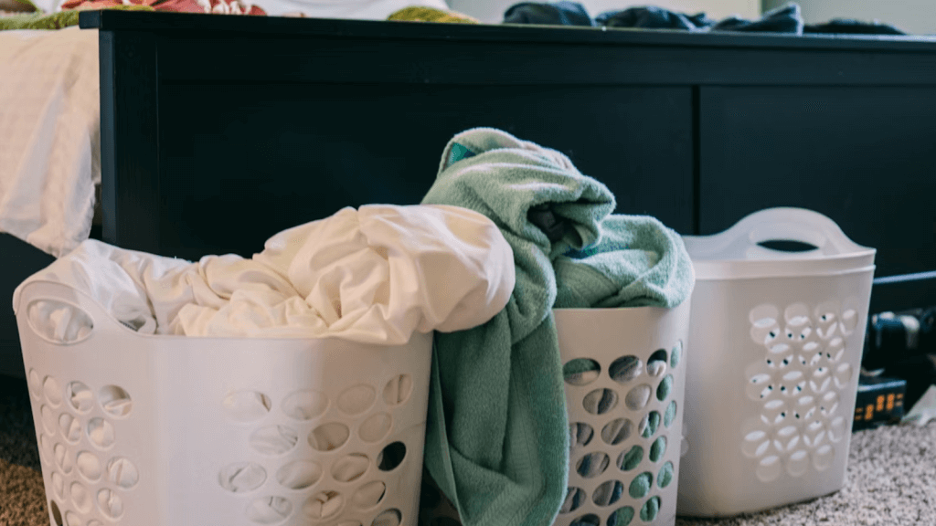 Three white laundry baskets filled with towels and bedding placed on a carpeted floor at the end of a bed.