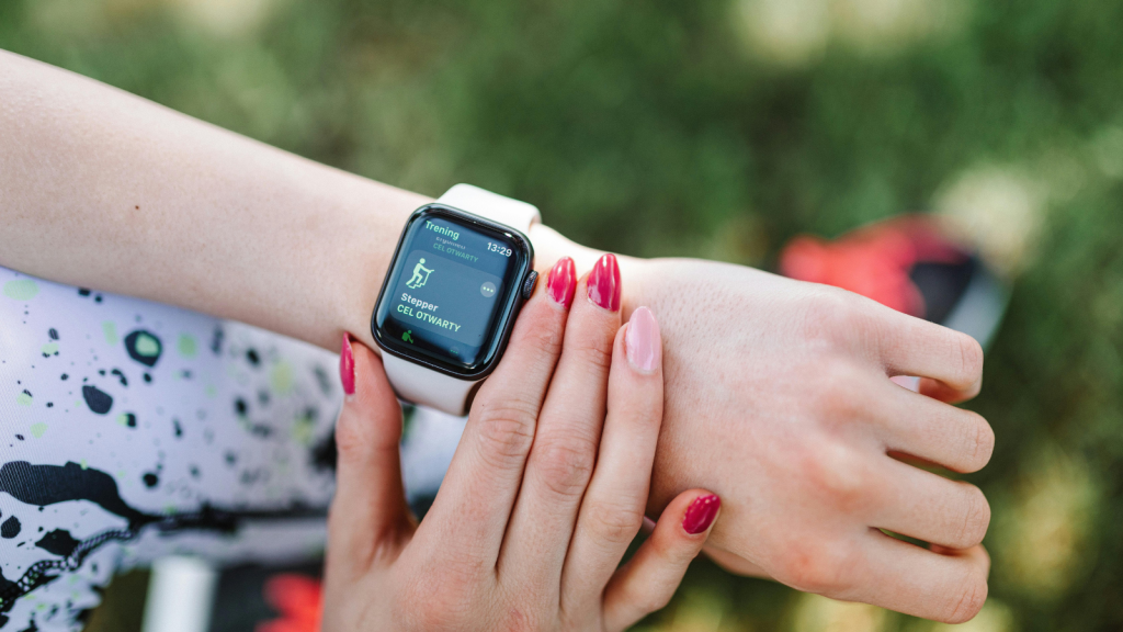 A person wearing patterned athletic leggings looks at a smartwatch showing a workout screen labeled “Trening” and “Stepper.” The person uses their other hand to interact with the watch; their nails are painted in alternating red and pink.