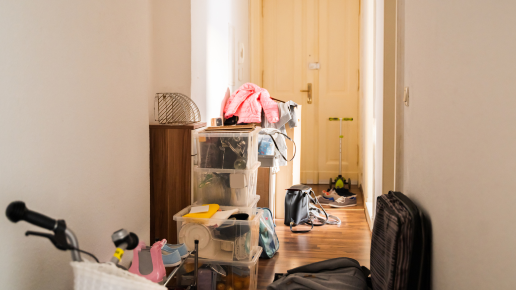 A narrow hallway with plastic bins, shoes, jackets, and scattered belongings leading toward a bright doorway.
