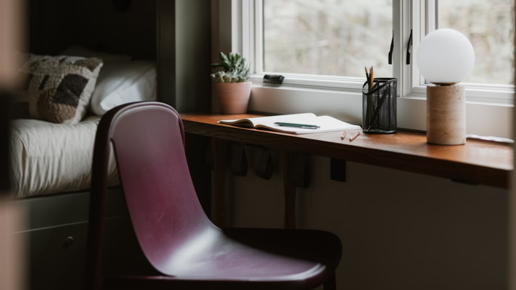 A simple wooden desk with a chair, open notebook, and pen beside a window with natural light and a potted plant.