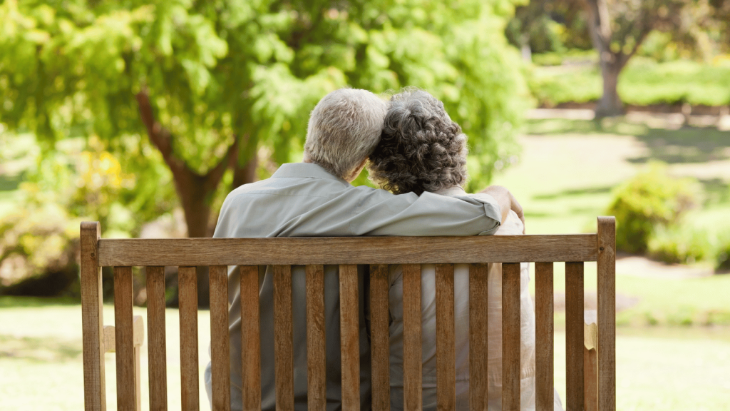 An older couple sits on a wooden park bench, the man’s arm around the woman, both looking out at a sunlit green landscape.