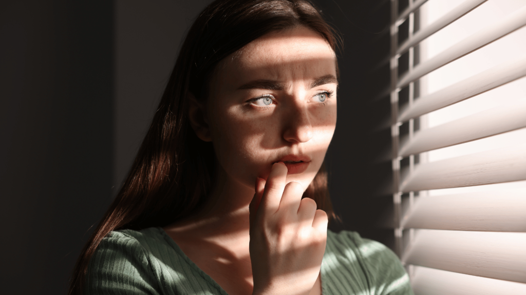 A young woman stands indoors near window blinds, her face partially covered by striped shadows from the light. She looks off to the side with a worried or contemplative expression, gently touching her lips with her fingers.