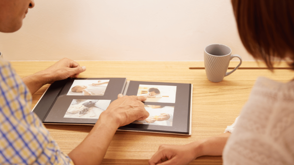Two people sit at a wooden table looking at a photo album. One person points to a picture of a baby while other infant photos appear on the open pages. A mug sits nearby.