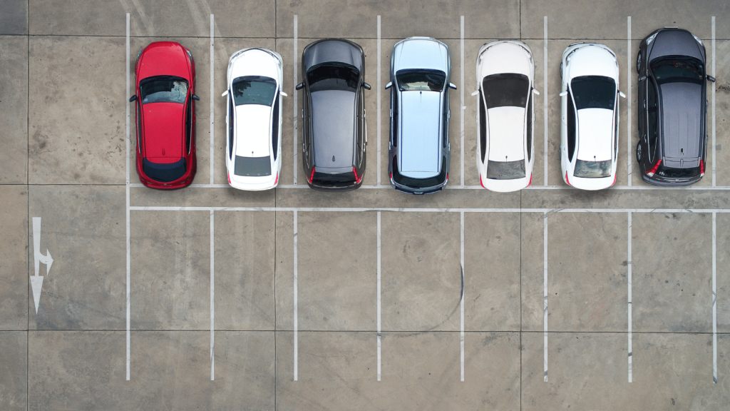 An aerial view of a mostly empty parking lot with a row of seven parked cars of various colors forming a neat line. The concrete surface and parking lines dominate the frame.