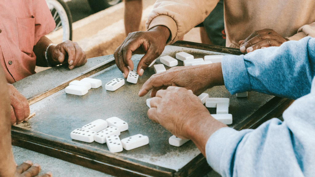 Close-up of several people’s hands as they play a game of dominoes on an outdoor table. White domino tiles are scattered across the surface.