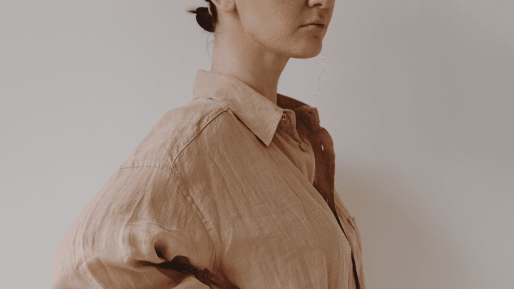 A close-up side view of a woman wearing a loose, wrinkled, light brown button-up shirt with her hair tied back. The background is simple and minimal.
