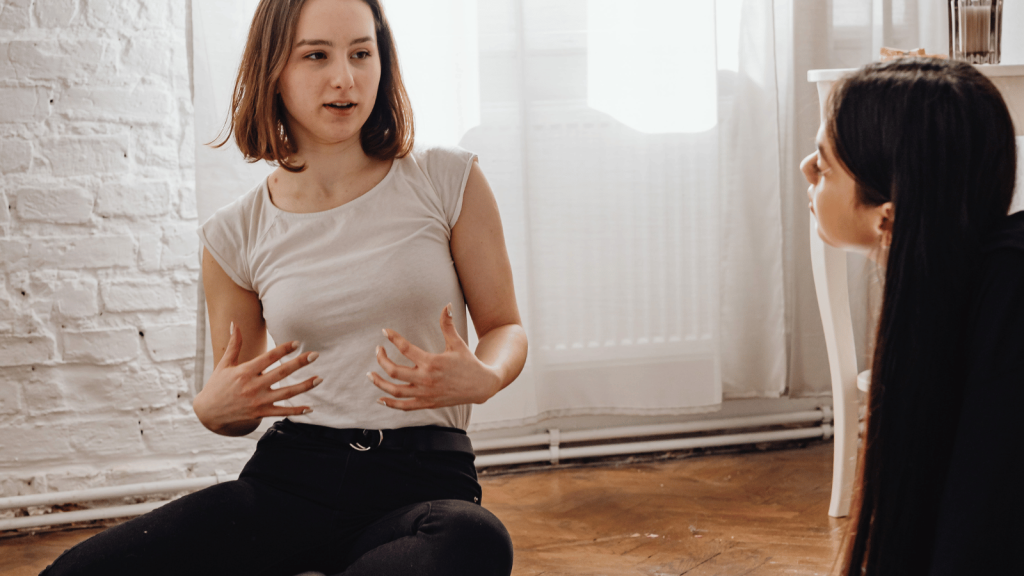 Two young women sit on a wooden floor in a bright room. One gestures with both hands while speaking, and the other listens attentively, leaning slightly forward. They appear engaged in a focused conversation.