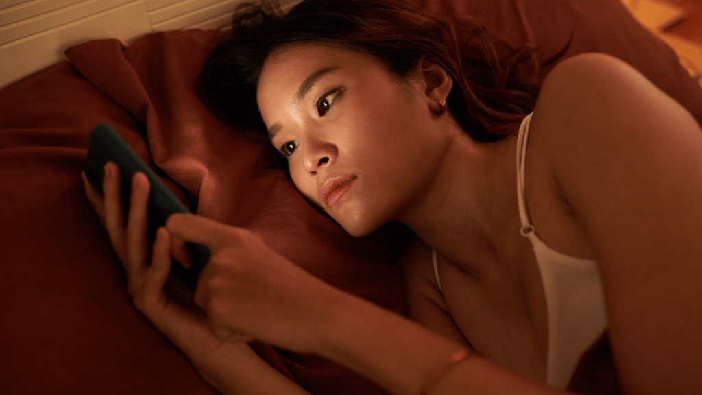 A woman lies in bed at night, illuminated by the glow of her phone as she scrolls with a blank, absorbed expression. Warm orange lighting casts shadows on the pillows and sheets around her.