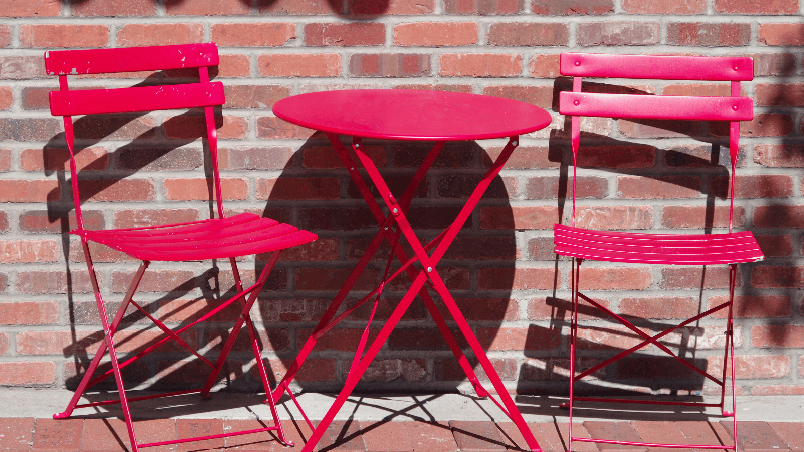 Two bright red metal chairs and a matching round table sit against a red brick wall in direct sunlight. The furniture casts crisp shadows on the bricks.