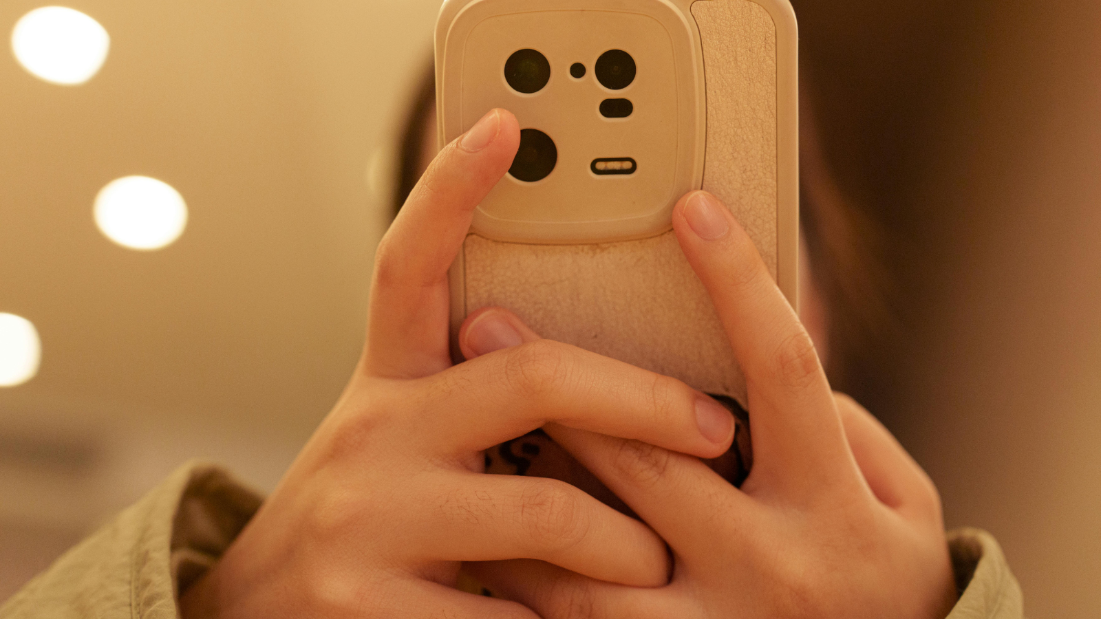 A close-up of hands holding a beige smartphone with a large camera module. The background features warm indoor lighting with circular bokeh from ceiling lights.