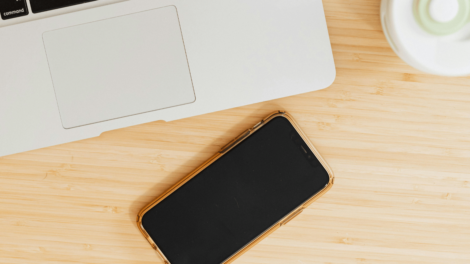 Featured A smartphone with a black screen rests on a wooden surface next to an open laptop and a white cup. The phone is centered in the frame, suggesting a workspace or casual desk environment.