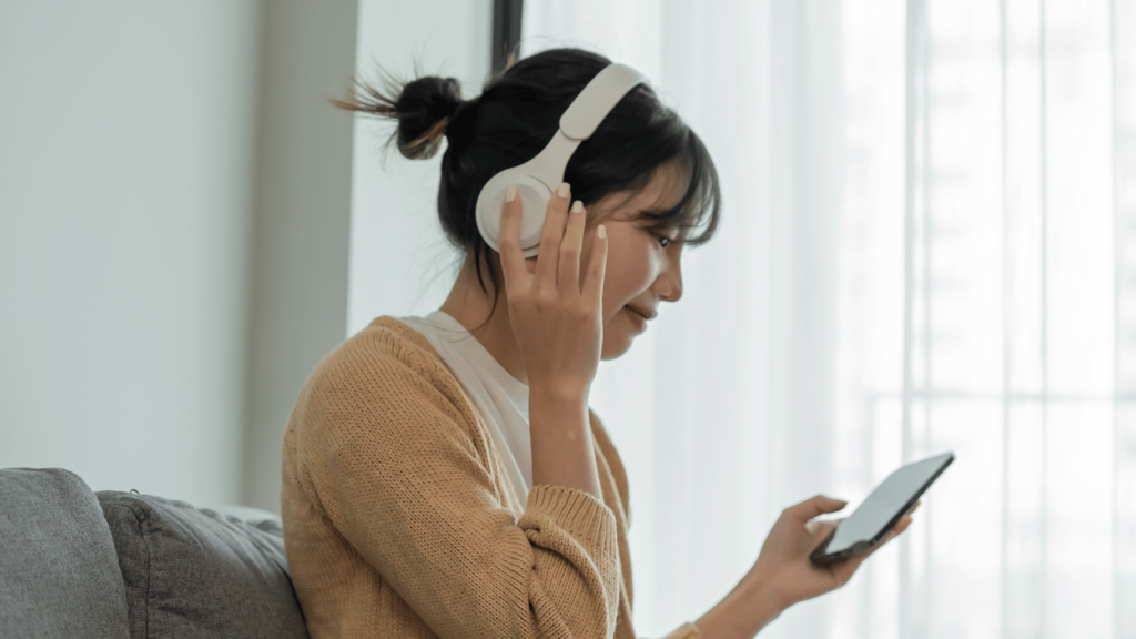 A woman wearing white over-ear headphones sits on a couch while looking at a smartphone. She holds one earcup with her hand as though adjusting or listening closely. Soft natural light comes through the window behind her.