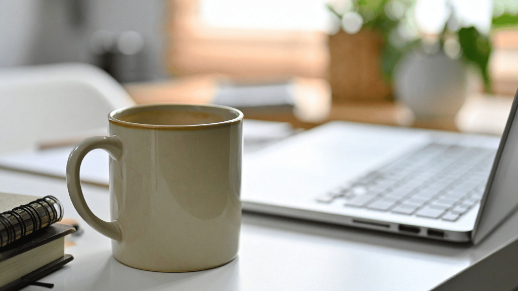 A beige coffee mug sits on a clean white desk next to a notebook and an open laptop. Soft natural light and blurred plants in the background create a calm workspace atmosphere.