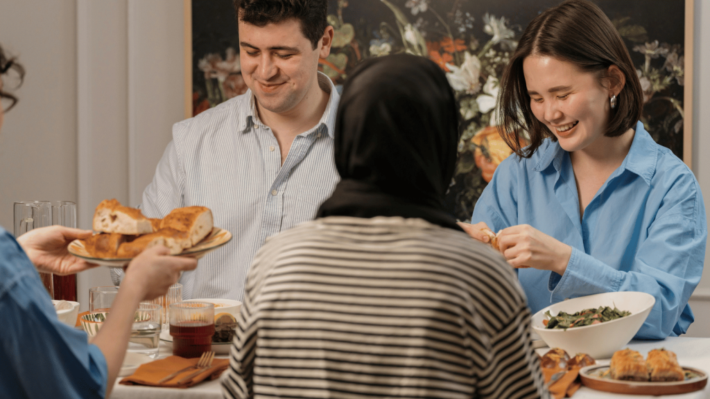 A group of people sit around a dining table sharing bread and food. Two smiling people face the camera while another, wearing a headscarf, sits with their back to the viewer. The scene is warm and communal.