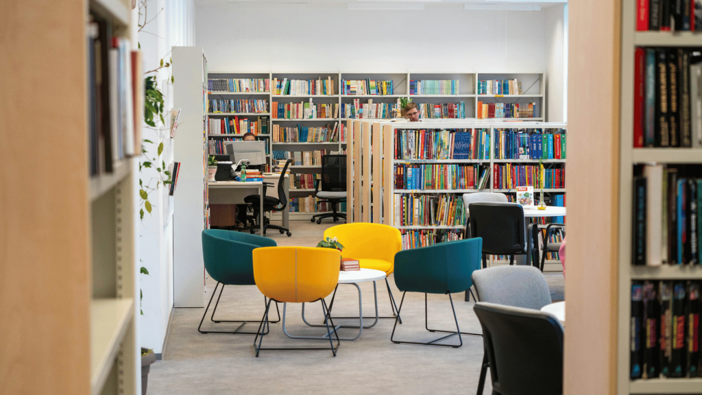 A bright, modern library interior featuring rows of bookshelves, several desks with computers, and a central seating area with colorful chairs around small round tables. A few people work at computers in the background. The space is clean, organized, and well lit.