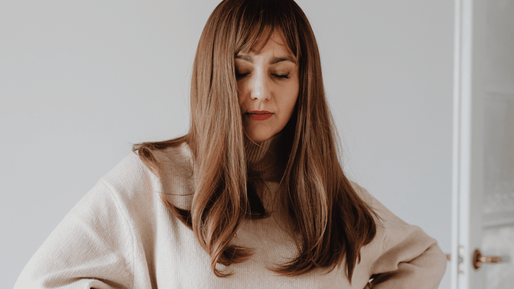 A close-up portrait of a woman with long brown hair wearing a beige turtleneck sweater. Her eyes are closed, and her hair falls partly over her face. The background is softly out of focus.