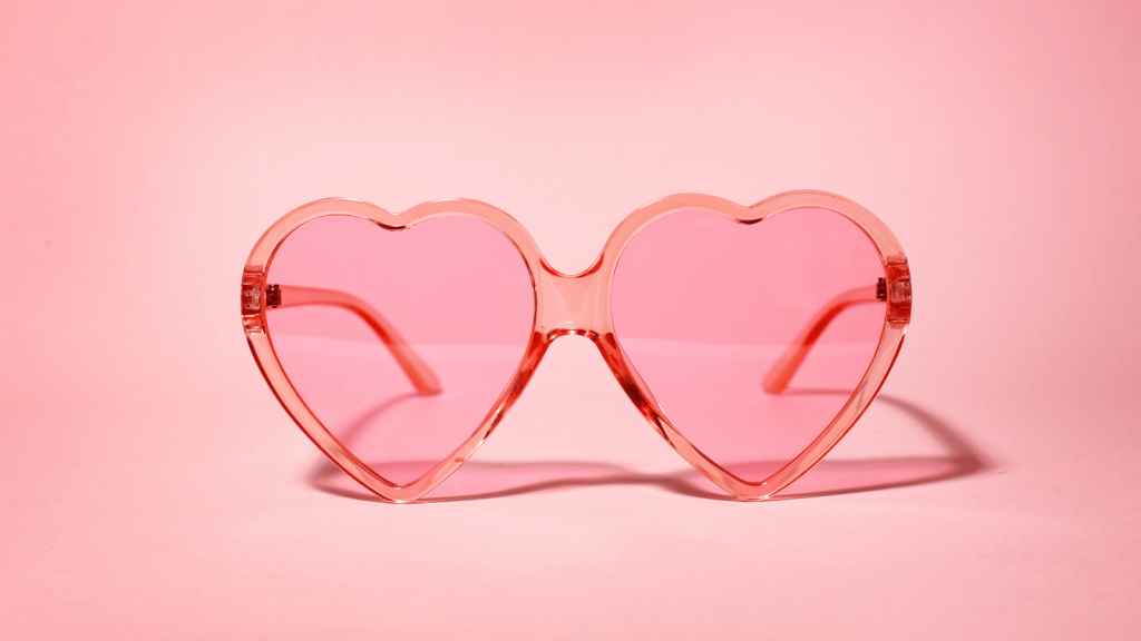 Close-up of pink, heart-shaped sunglasses with translucent frames and lenses, set against a solid pink background.