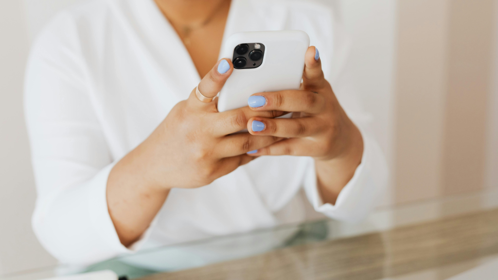 A person with light-blue nail polish holds a white smartphone with both hands. The person is wearing a white long-sleeve top, and the background is softly blurred.