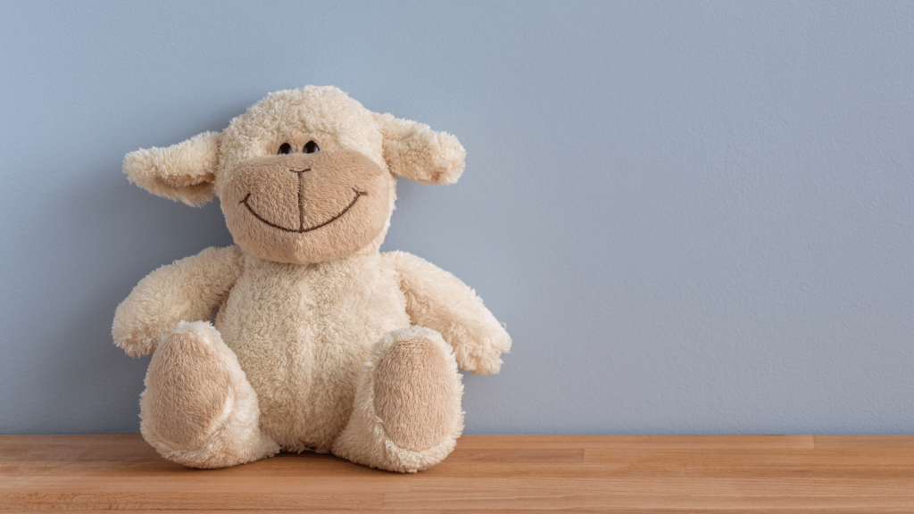 A cream-colored stuffed lamb with a stitched smile sits on a wooden surface against a pale blue wall.
