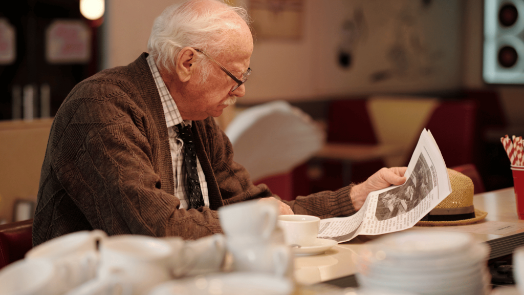 An older man with white hair and glasses sits alone in a café-style setting, reading a newspaper while holding a cup of coffee. Stacks of cups and a straw holder sit on the counter nearby. The atmosphere is warm and nostalgic.