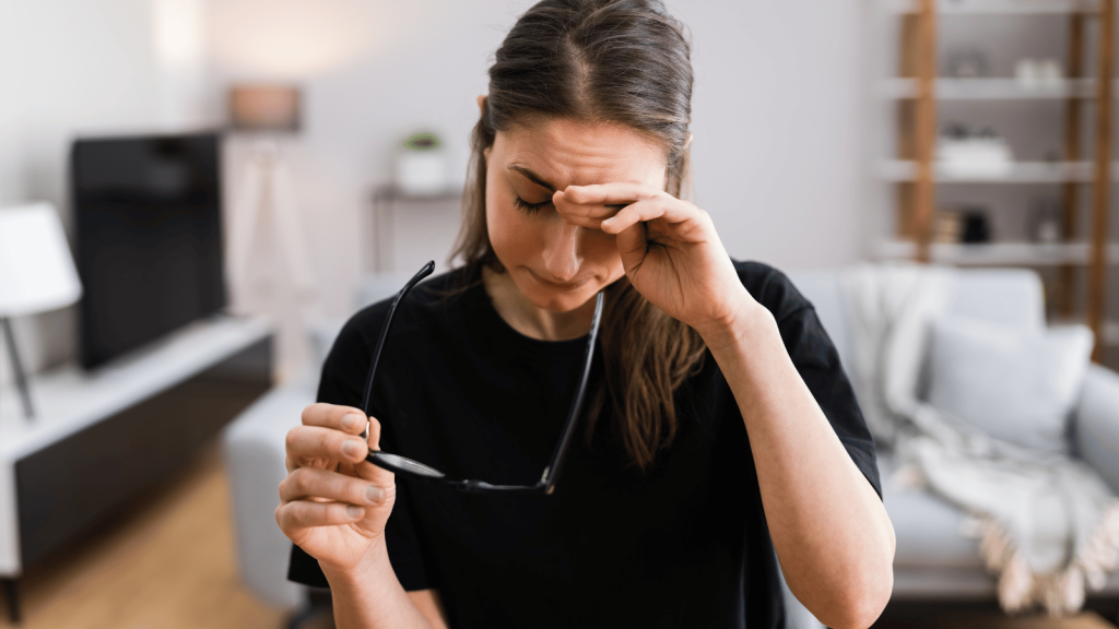 A woman in a black shirt stands in a bright living room, removing her glasses while rubbing her eyes with a strained expression. Shelves, plants, and soft décor appear in the background.