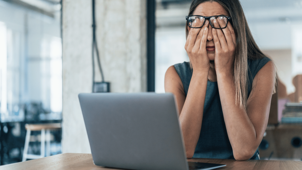 A woman sits at a desk with her laptop open, covering her eyes with both hands in exhaustion. She wears glasses and a sleeveless top, and the workspace around her looks industrial and modern.