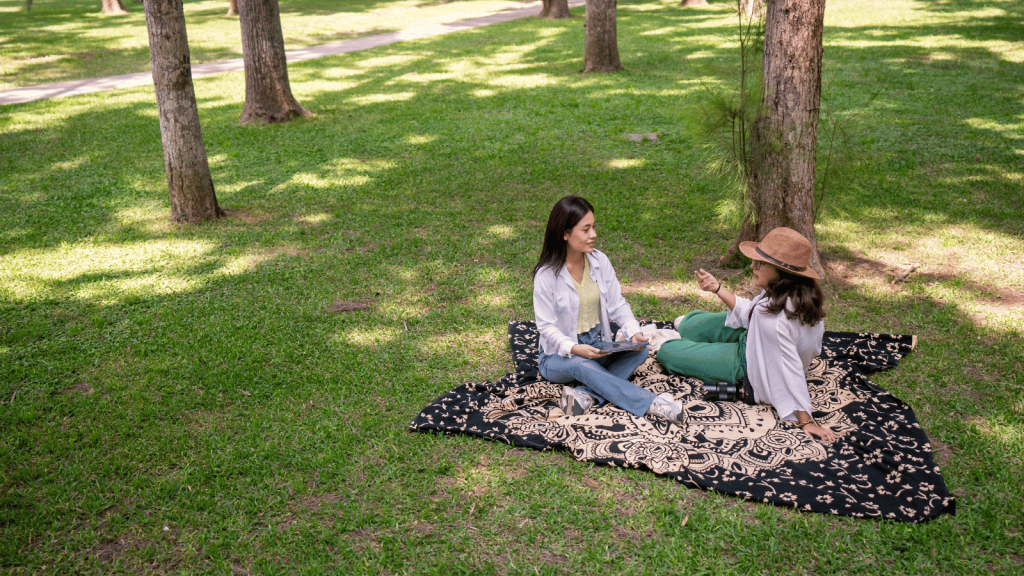 Two women sit on a patterned picnic blanket in a grassy park, talking and smiling. One holds a notebook, and the other gestures as she speaks. Trees cast dappled shadows across the lawn.