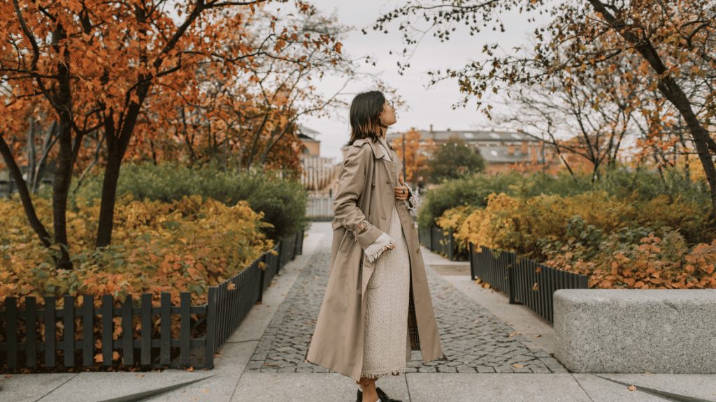 A woman walking through a landscaped urban park with autumn foliage. She wears a beige trench coat over a textured cream dress and looks upward as she walks along a stone path lined with plants and small fences.